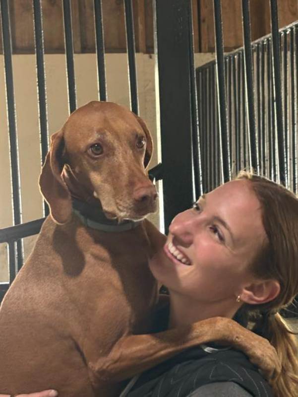 Smiling woman hugging a brown dog inside stable.